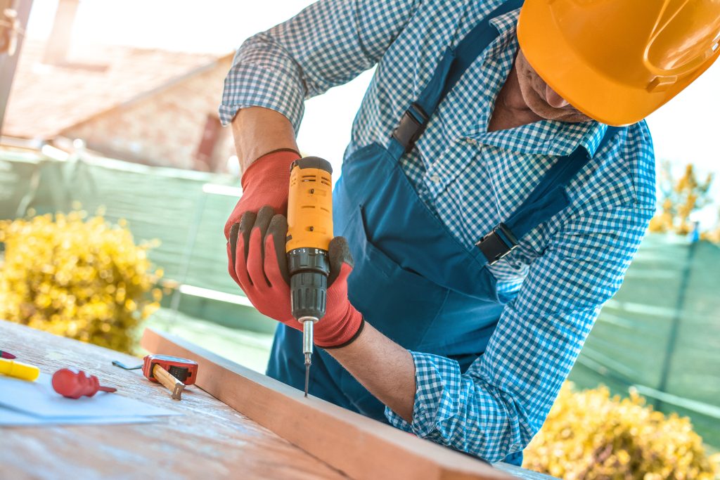 Hand of worker drills a hole with wooden plank using electric drill machine in workshop